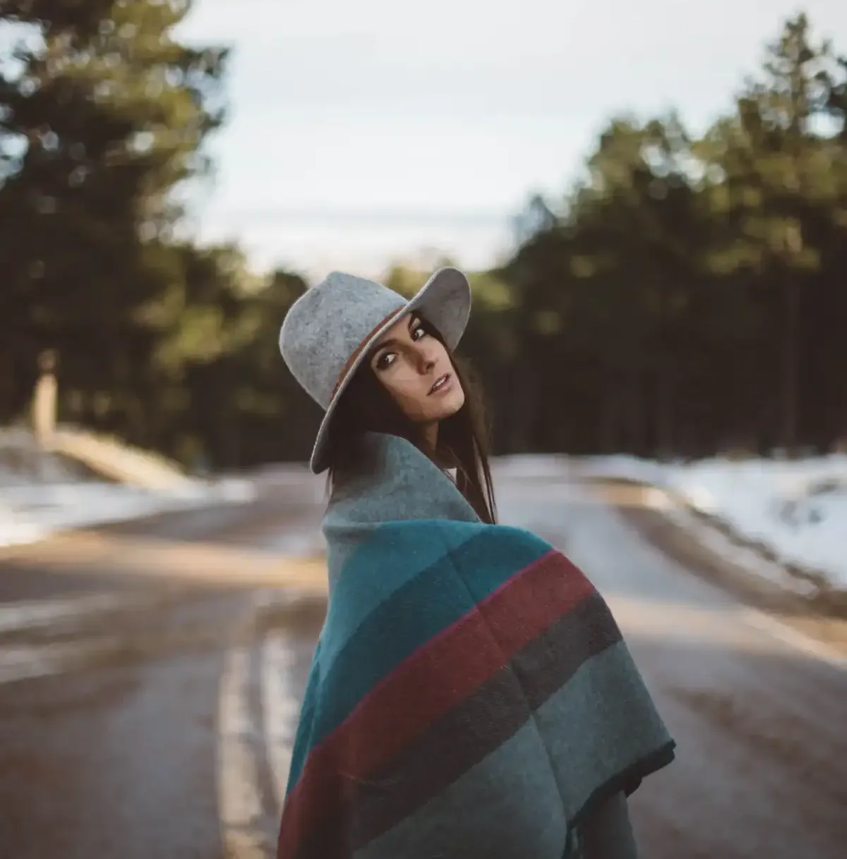 Woman with wide-brimmed hat and warm blanket standing on a snowy forest road during winter.